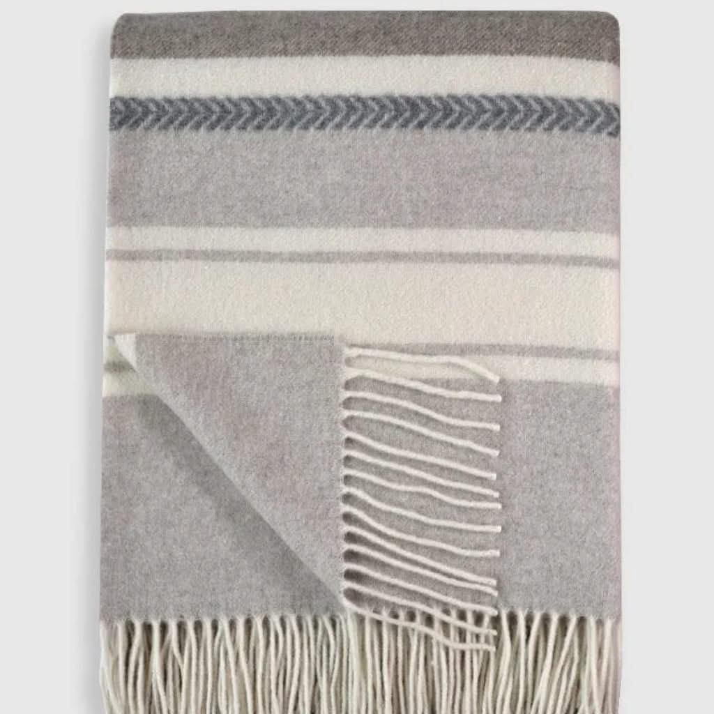 Folded Muskoka Throw in merino wool and cashmere blend with gray and cream stripes, featuring fringe detail. Ideal for luxury bedding or gifting.