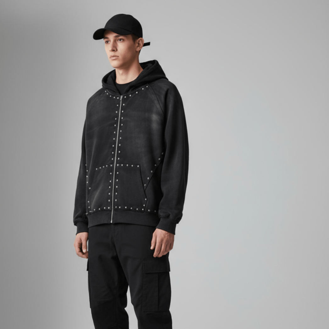 Male model wearing unisex black studded zip-up hoodie styled with black cargo pants. Rugged streetwear look with vintage cotton texture.