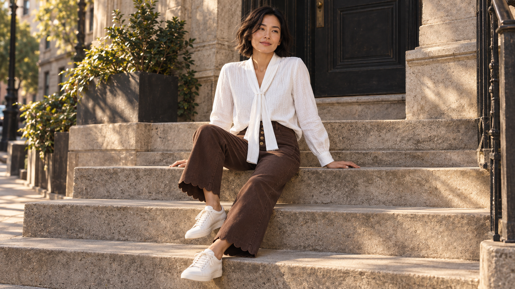 Woman seated on city steps wearing the white scarf-tie blouse and brown scalloped-hem jeans